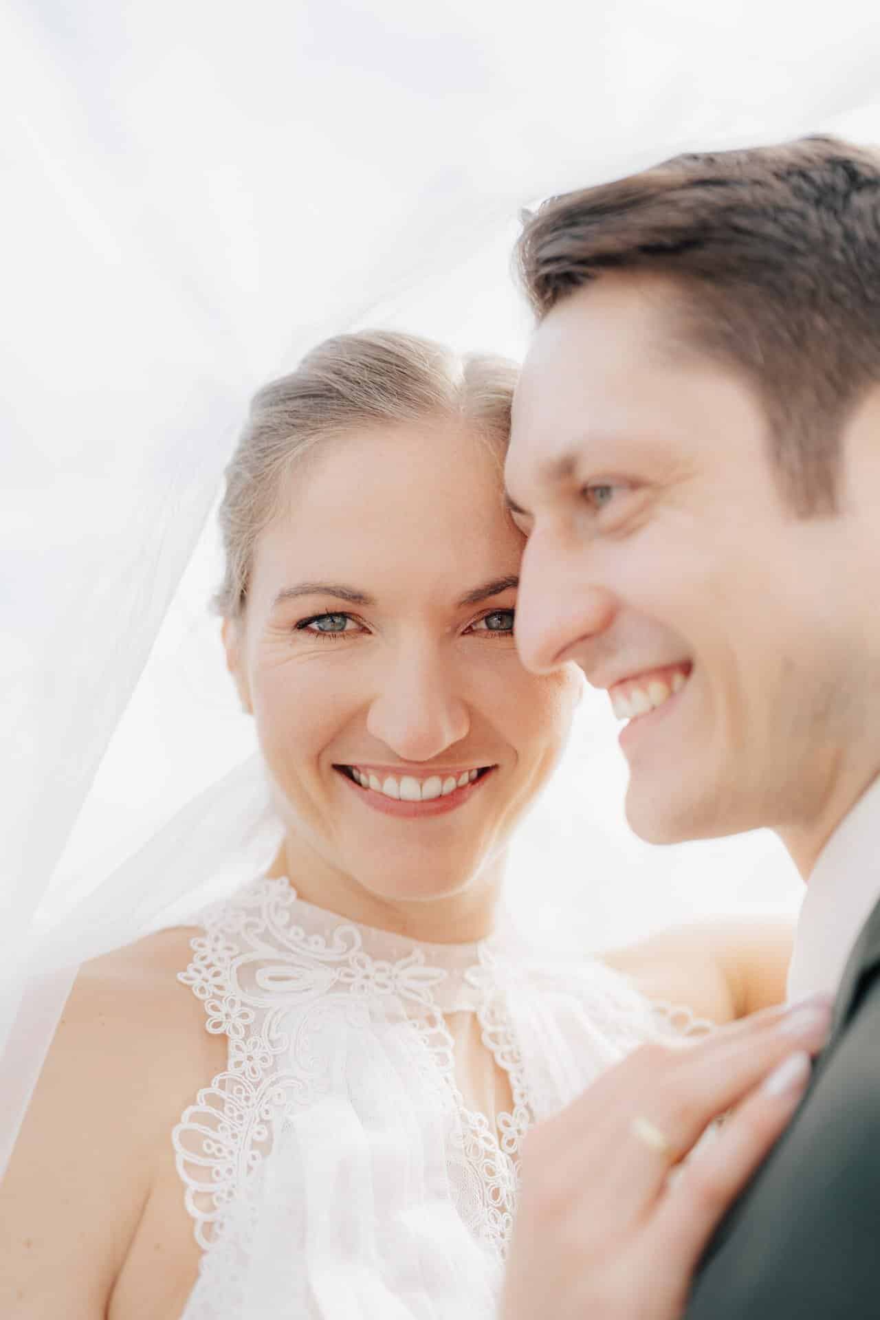 Eine Nahaufnahme eines lächelnden Brautpaares an ihrem Hochzeitstag. Die von Hochzeitsfotograf Göppingen aufgenommene Braut trägt ein weißes Spitzenkleid und einen Schleier, während der Bräutigam einen Anzug trägt. Beide sehen glücklich aus und genießen den gemeinsamen Moment.