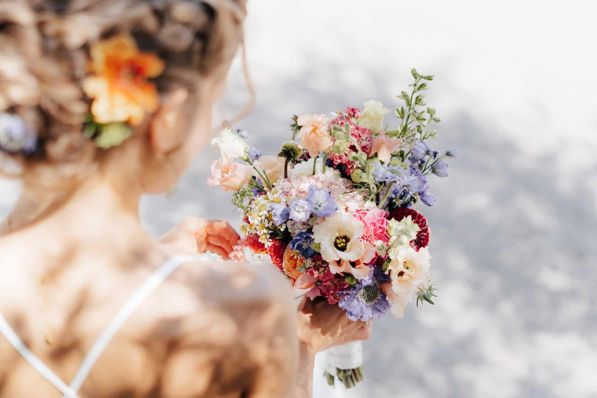 Eine Frau mit Blumen in ihrem lockigen Haar hält einen bunten Blumenstrauß an einem hellen, sonnigen Tag. Aufgenommen von einem Hochzeitsfotograf Göppingen, trägt sie ein weißes Kleid und ist von hinten inmitten von leuchtend rosa, lila, blauen und weißen Blüten zu sehen.