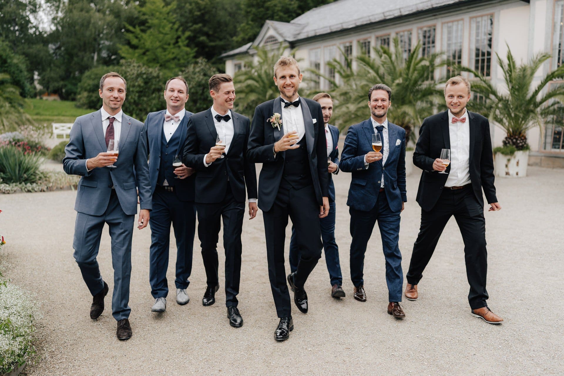 Eine Gruppe von sieben Männern in Anzügen, lächelnd und mit Getränken in der Hand, spazieren gemeinsam im Freien in der Nähe eines großen Glasgebäudes mit üppigem Grün und Palmen im Hintergrund - perfekt eingefangen von einem Hochzeitsfotograf Göppingen.