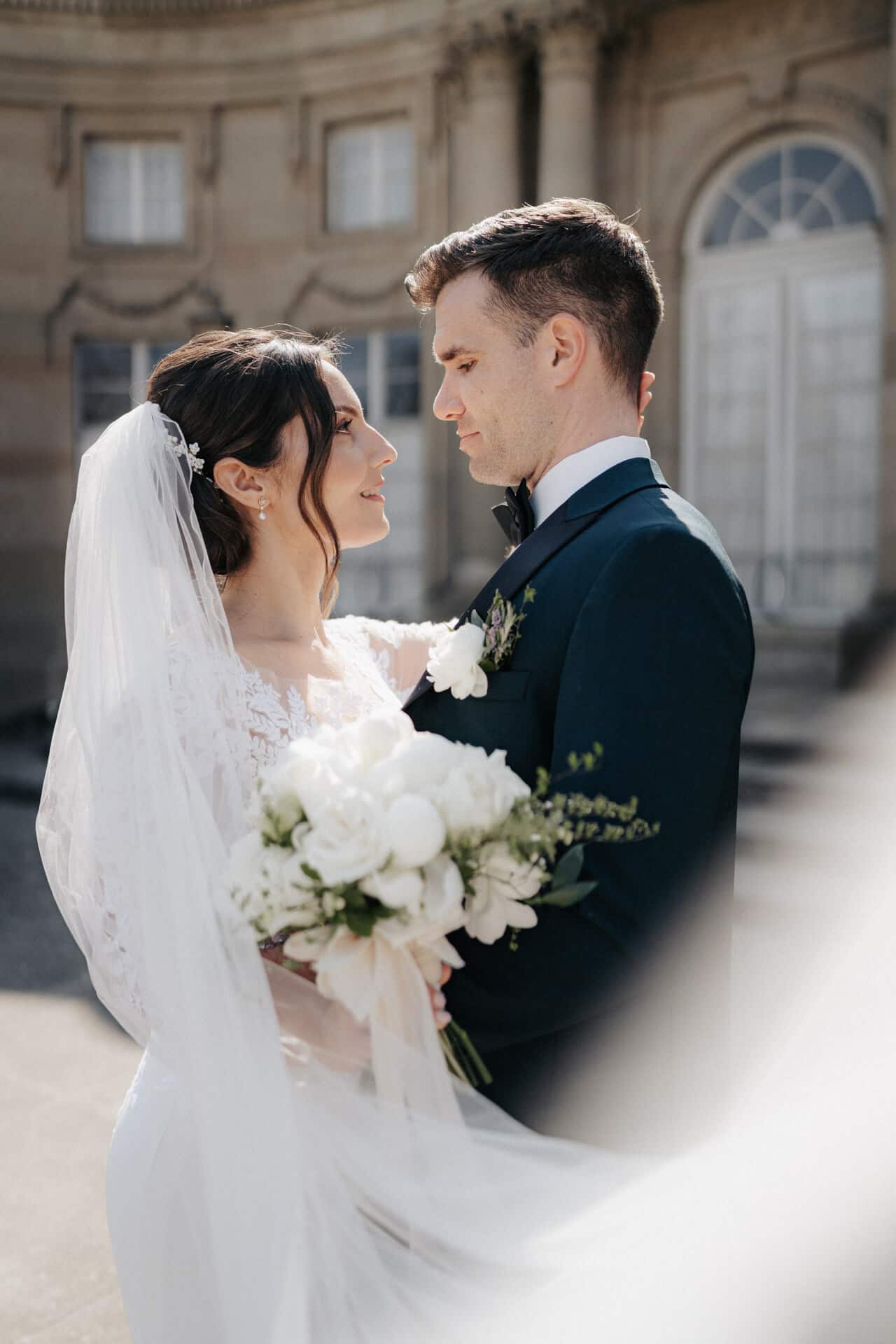 Eine Braut und ein Bräutigam stehen im Freien dicht beieinander und blicken sich in die Augen. Die Braut hält einen Strauß weißer Blumen in der Hand, der Bräutigam trägt einen dunklen Anzug, im Hintergrund ist ein historisches Gebäude zu sehen.