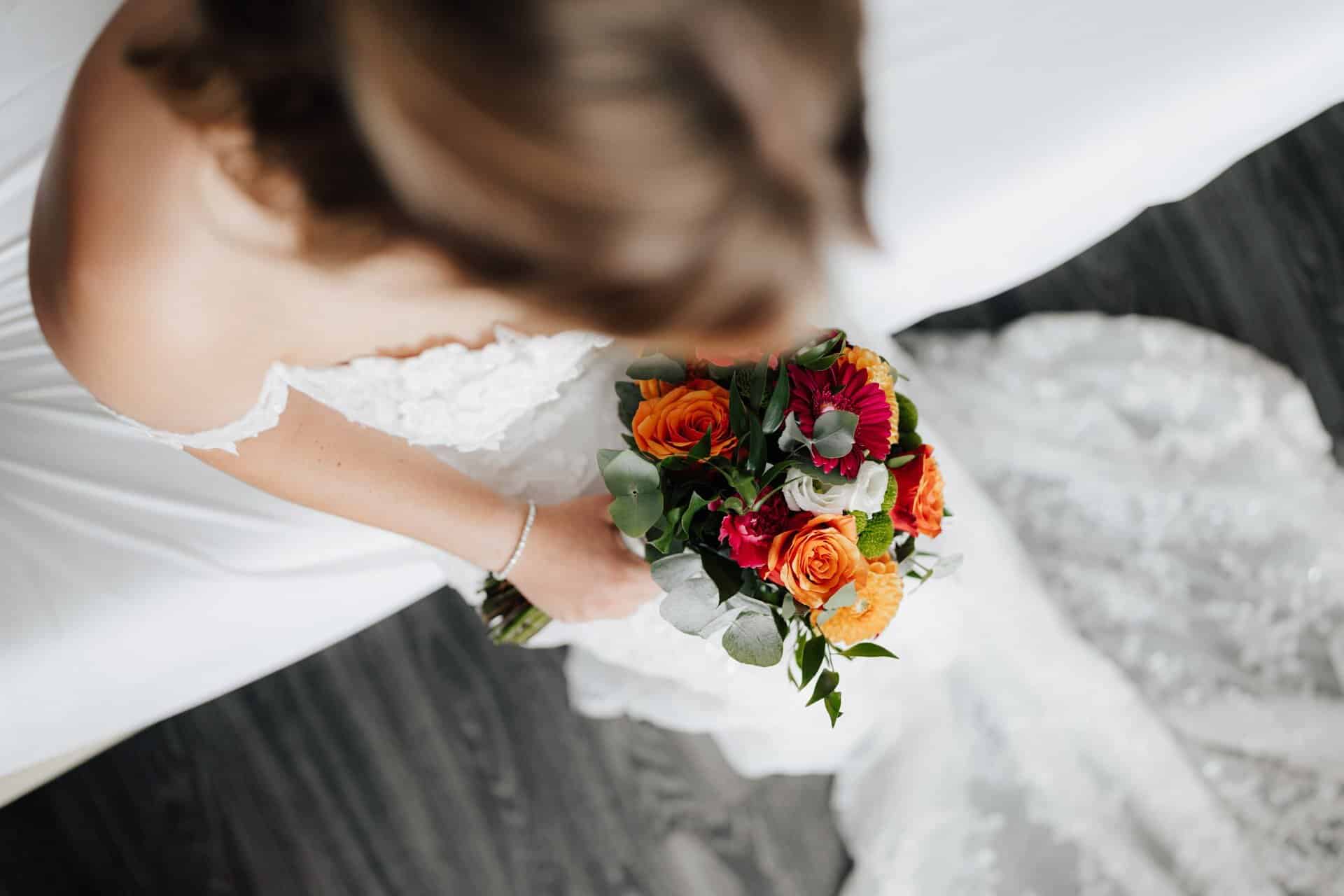 Eine Braut in einem weißen Hochzeitskleid sitzt auf einem Bett und hält einen Strauß aus orangefarbenen und rosafarbenen Blumen mit Grünzeug. Von einem Hochzeitsfotograf Göppingen von oben aufgenommen, hebt das Foto den Strauß hervor und enthüllt teilweise ihr Gesicht.