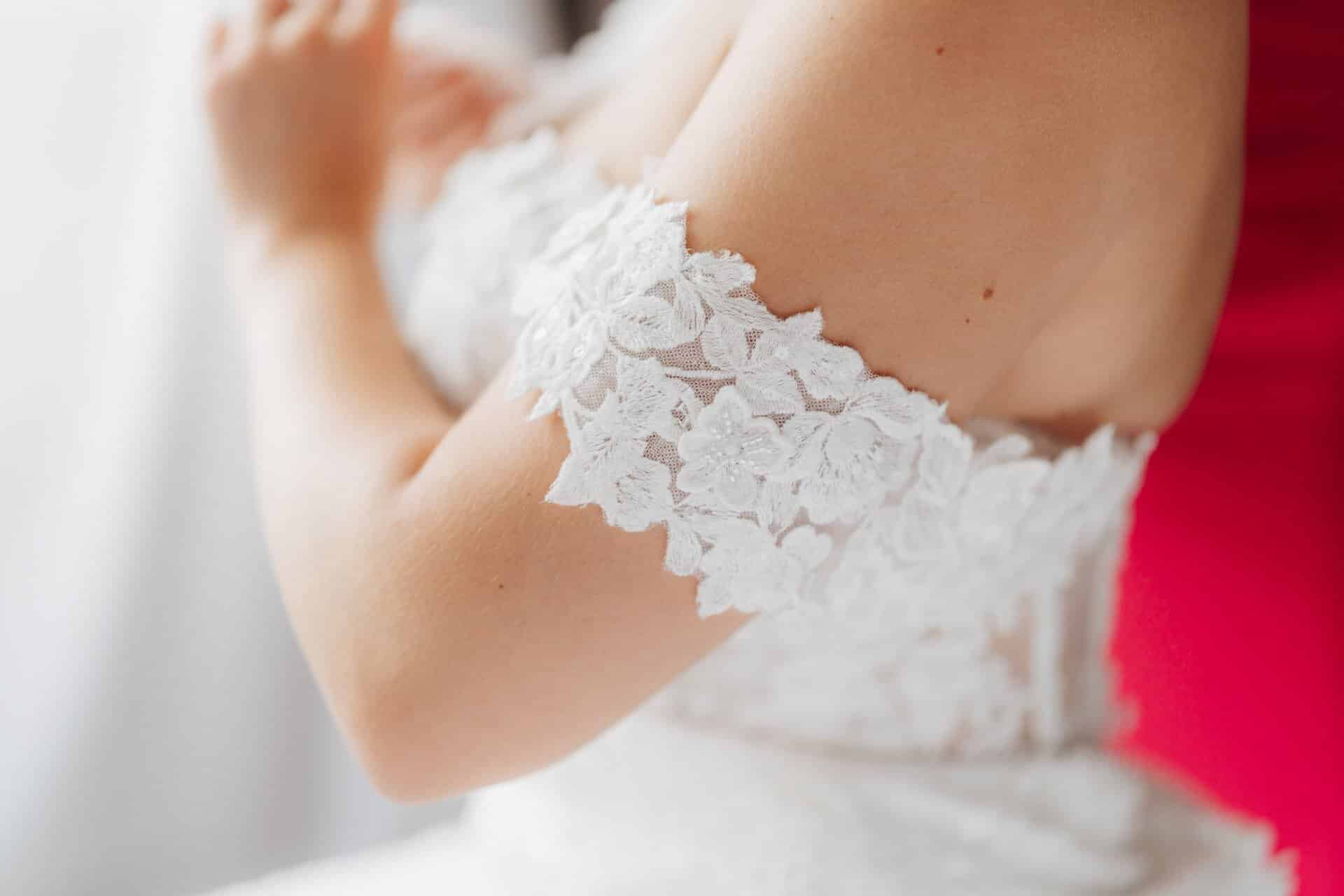 Eine Nahaufnahme einer Person, die ein schulterfreies weißes Spitzenhochzeitskleid trägt, aufgenommen von Hochzeitsfotograf Göppingen, wobei der Schwerpunkt auf den zarten floralen Spitzendetails an den Ärmeln und am oberen Mieder liegt.