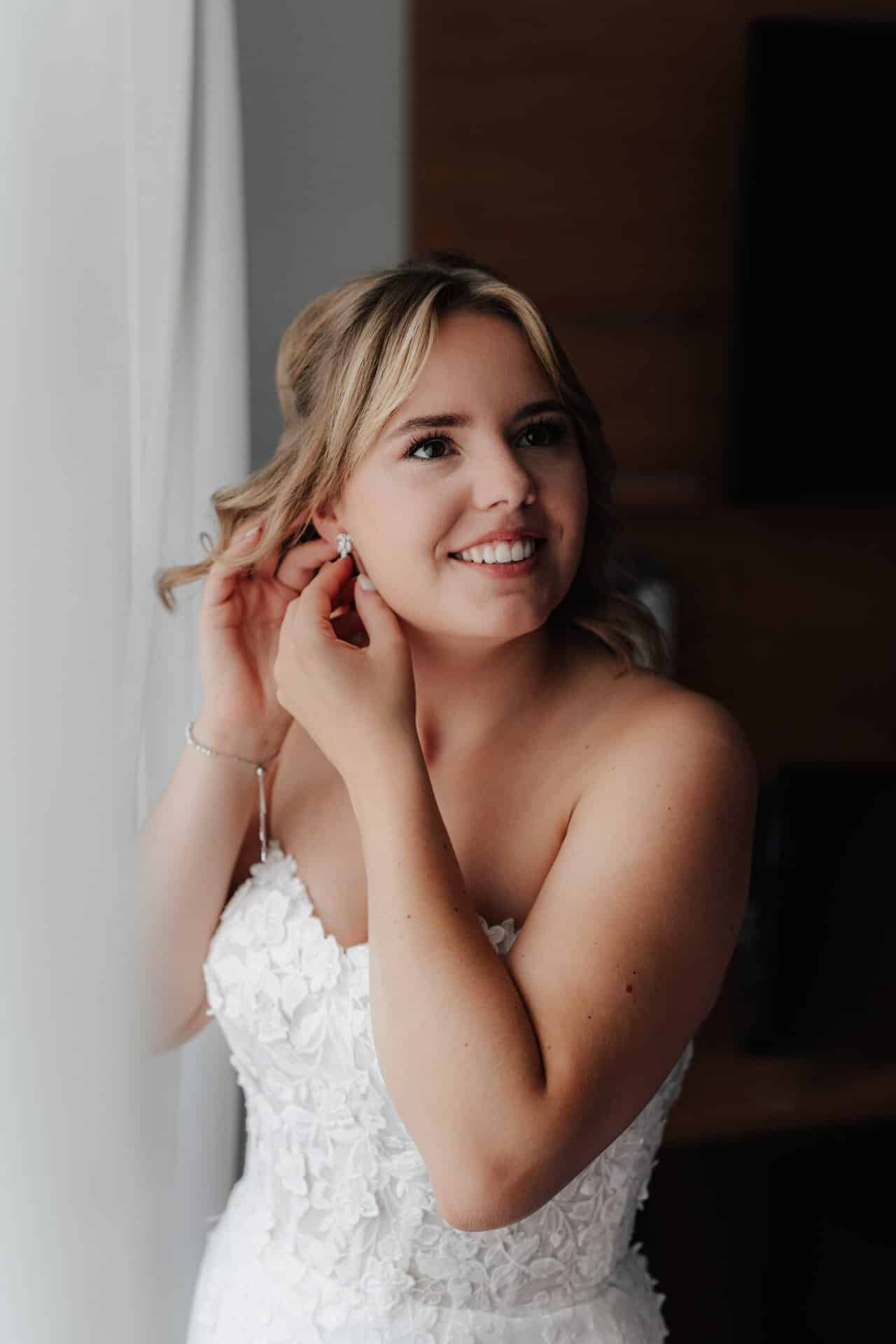 Eine lächelnde Braut in einem trägerlosen weißen Spitzenkleid steht an einem Fenster und rückt ihren Ohrring zurecht, während sanftes natürliches Licht ihr Gesicht beleuchtet - wunderschön eingefangen von Hochzeitsfotograf Göppingen.