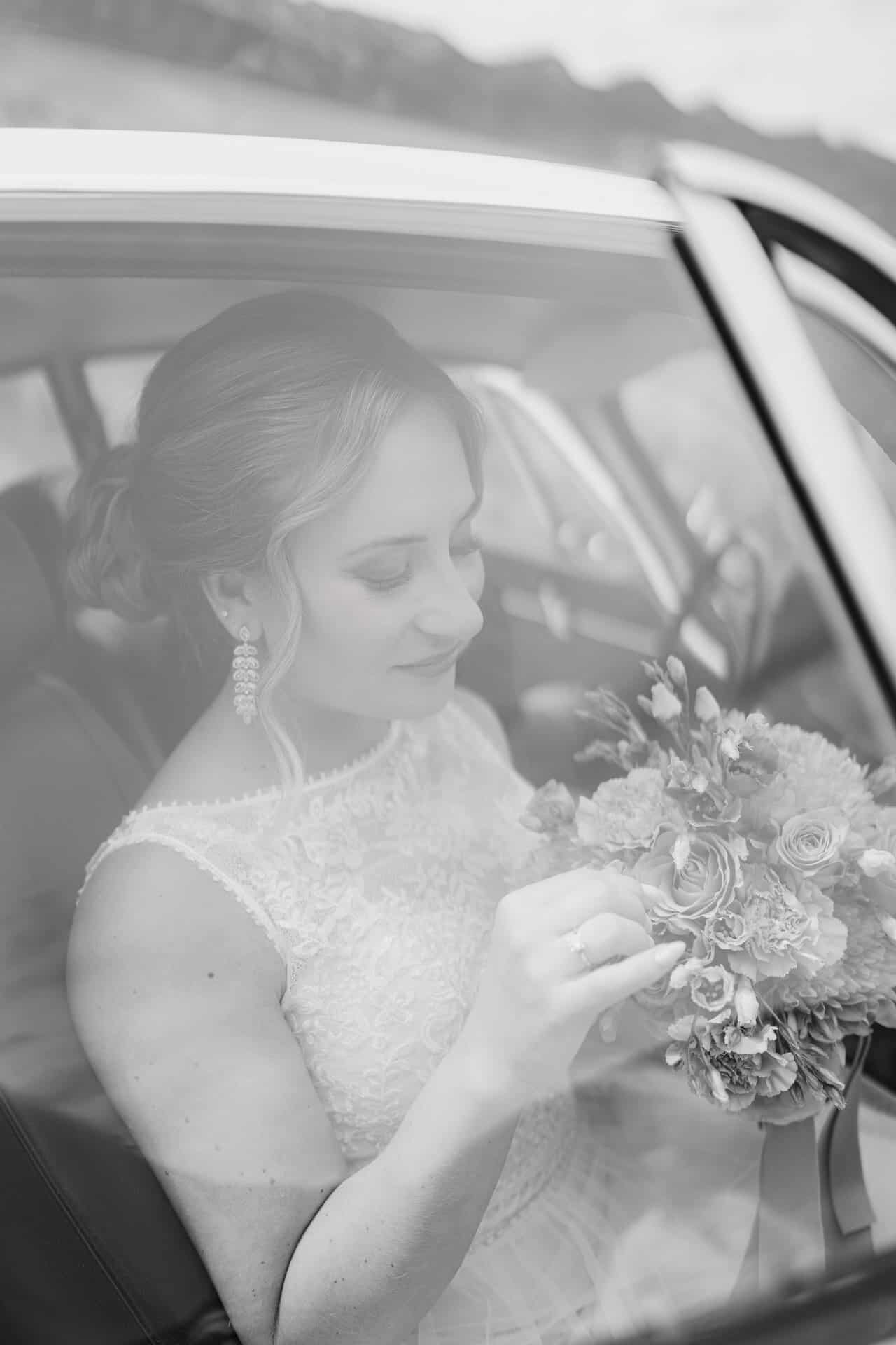 Eine Braut im Spitzenhochzeitskleid sitzt in einem Auto und bewundert ihren Blumenstrauß. Das Schwarz-Weiß-Foto wurde von einem Hochzeitsfotograf Göppingen aufgenommen und spiegelt ihre ruhige und nachdenkliche Stimmung wider.
