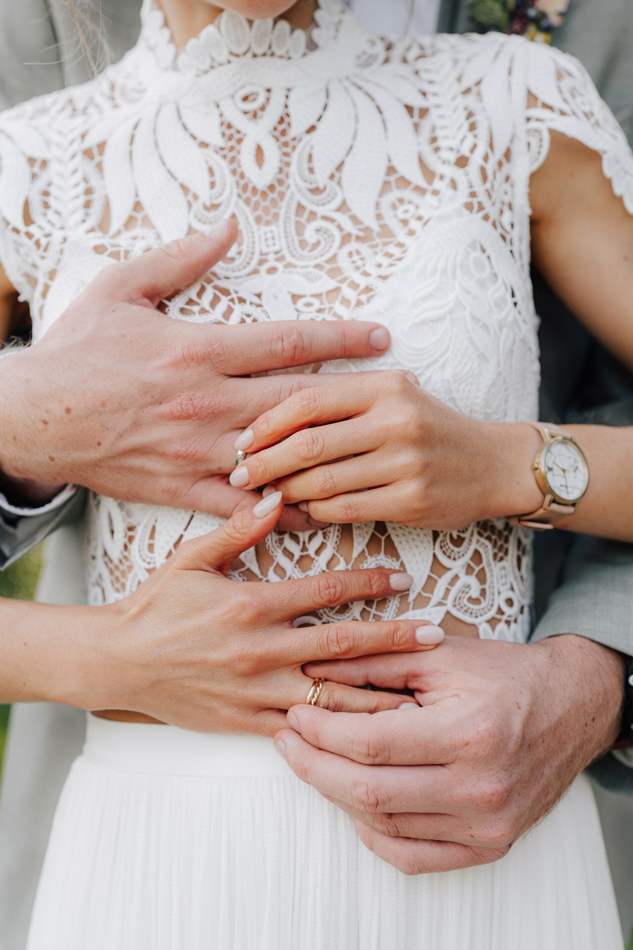 Eine Nahaufnahme von Hochzeitsfotograf Karlsruhe zeigt ein Paar in Hochzeitskleidung, das sich umarmt, wobei sich die Hände sanft um die Taille der Braut legen. Sie trägt ein Spitzenkleid, eine goldene Uhr und Ringe; die Hände des Bräutigams sind liebevoll hinter den ihren zu sehen.
