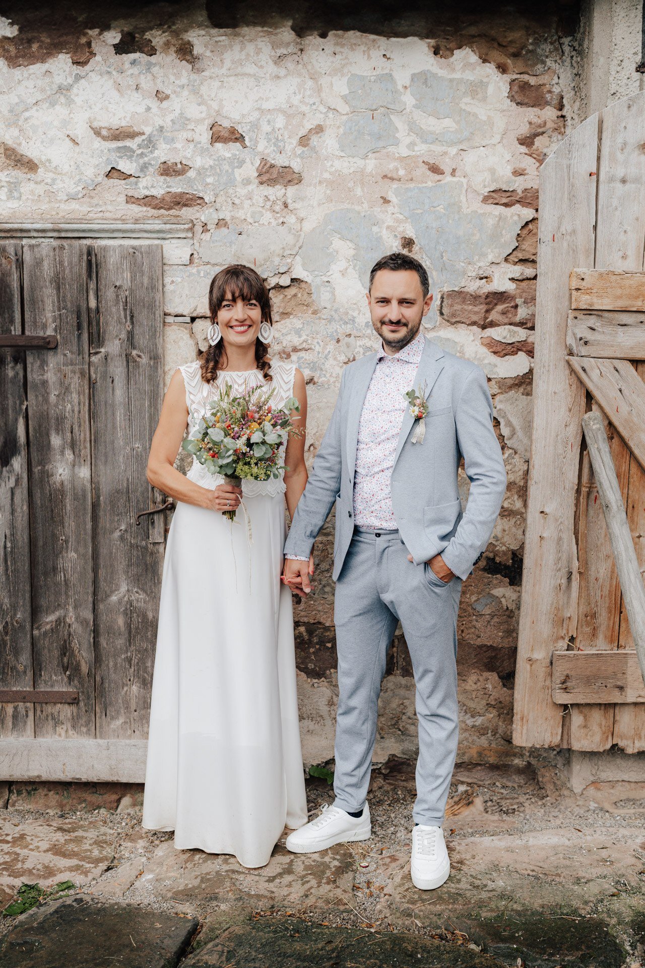 Eine Braut in einem weißen Kleid und ein Bräutigam in einem hellblauen Anzug stehen Händchen haltend vor einer rustikalen Steinmauer und Holztüren. Aufgenommen von einem Hochzeitsfotograf Karlsruhe, hält die Braut einen Blumenstrauß und beide lächeln leicht.