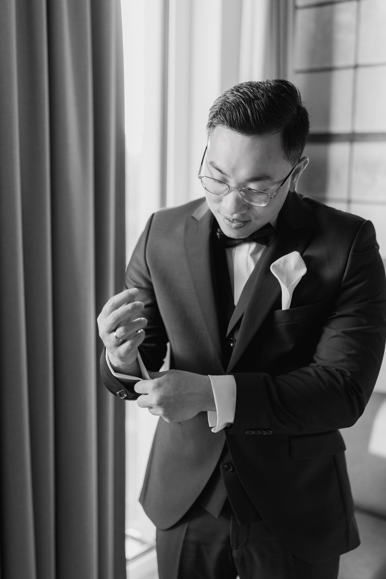 Ein Mann im Anzug und mit Fliege rückt seinen Manschettenknopf in der Nähe eines vorgehängten Fensters zurecht. Er trägt eine Brille und scheint sich auf eine formelle Veranstaltung vorzubereiten. Der Hochzeitsfotograf Ludwigsburg hat den Moment in Schwarz-Weiß festgehalten, der zeitlos wirkt.