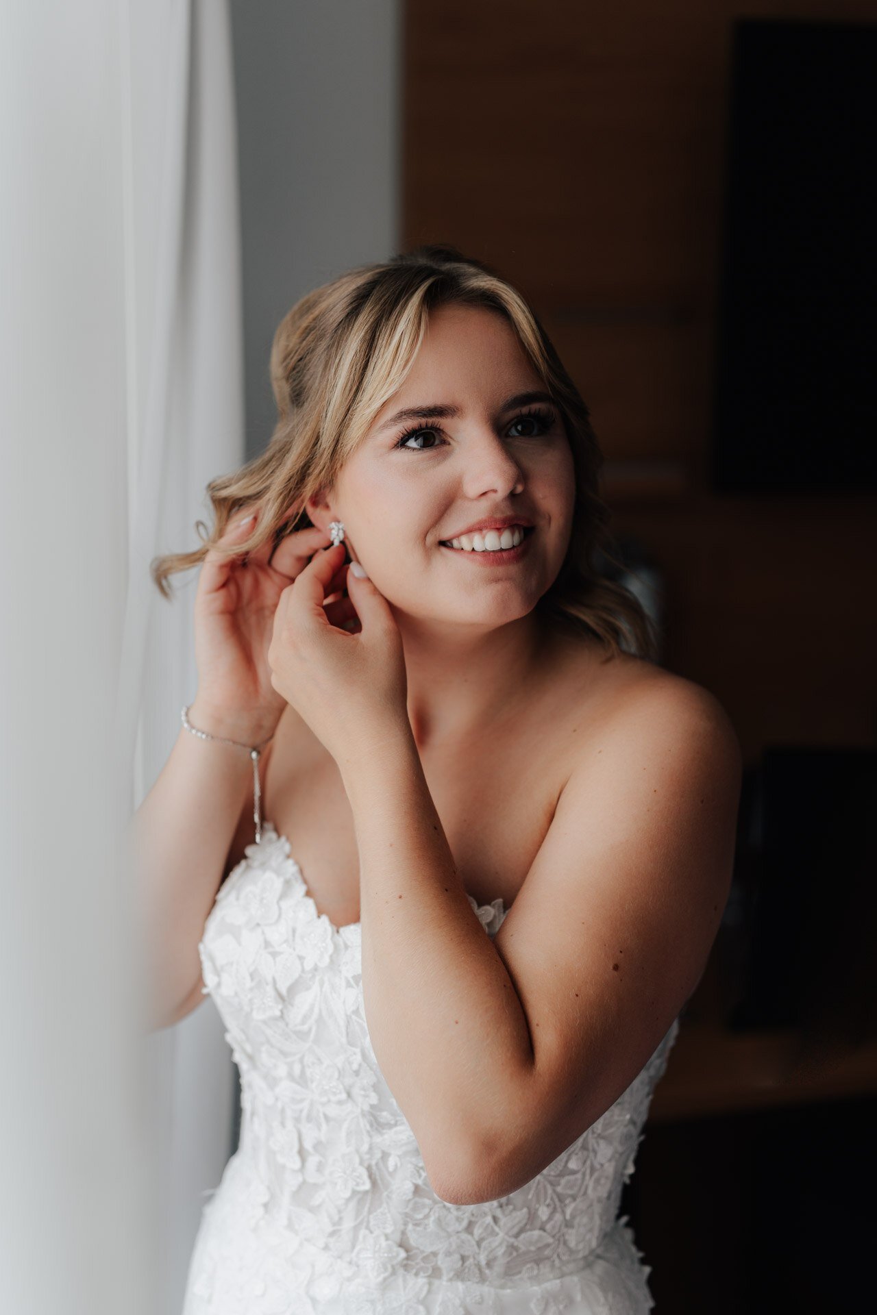 Eine lächelnde Braut in einem trägerlosen weißen Kleid mit Blumendetails steht an einem Fenster und steckt sich einen Ohrring an. Aufgenommen von einem Hochzeitsfotograf Ludwigsburg, beleuchtet natürliches Licht ihren glücklichen, heiteren Ausdruck, während sie nach draußen blickt.