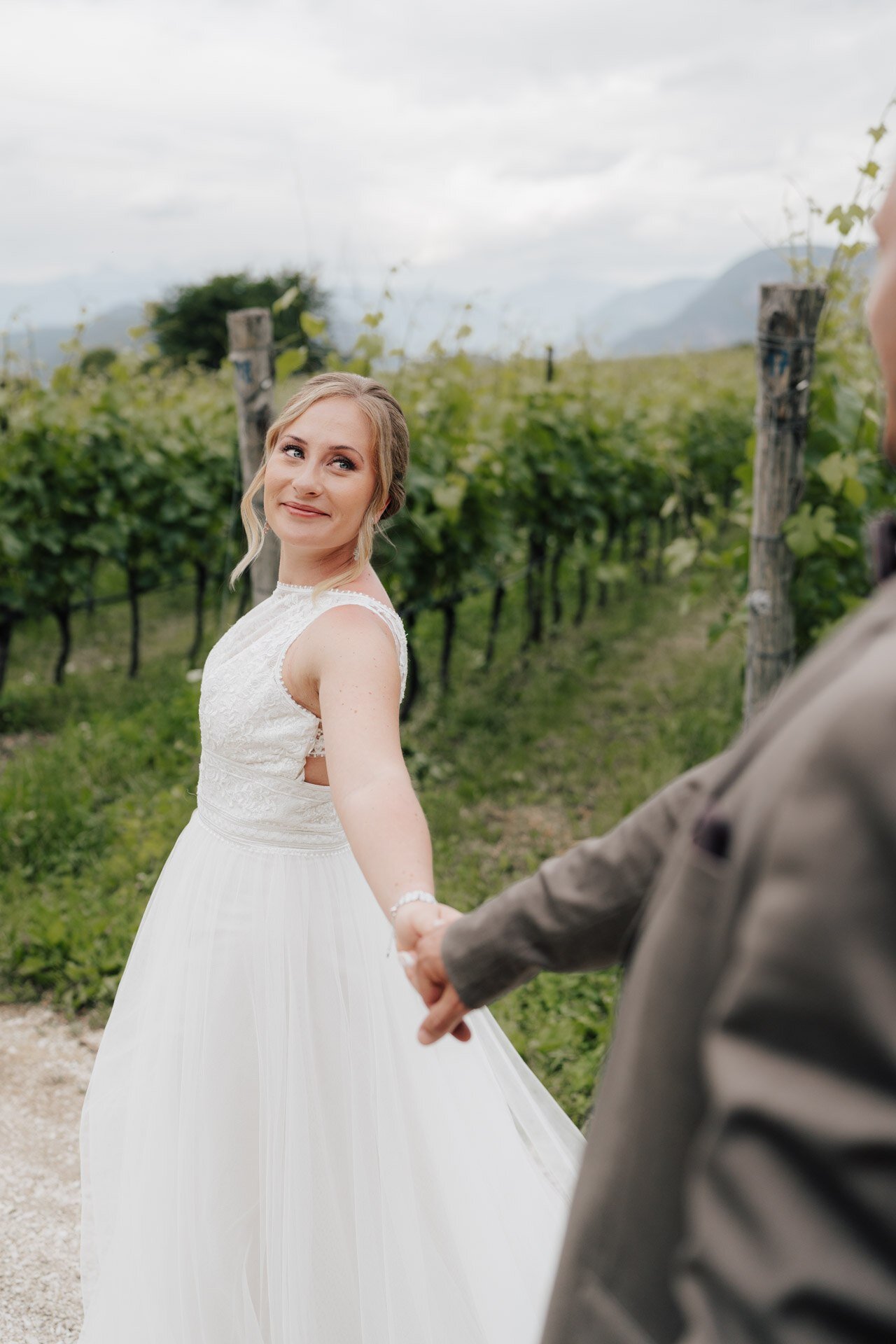 Eine Braut in einem weißen Kleid lächelt, während sie ihren Partner an der Hand hält und sie durch einen Weinberg mit grünen Reben und fernen Bergen führt, wunderschön eingefangen von einem Hochzeitsfotografen Ludwigsburg.