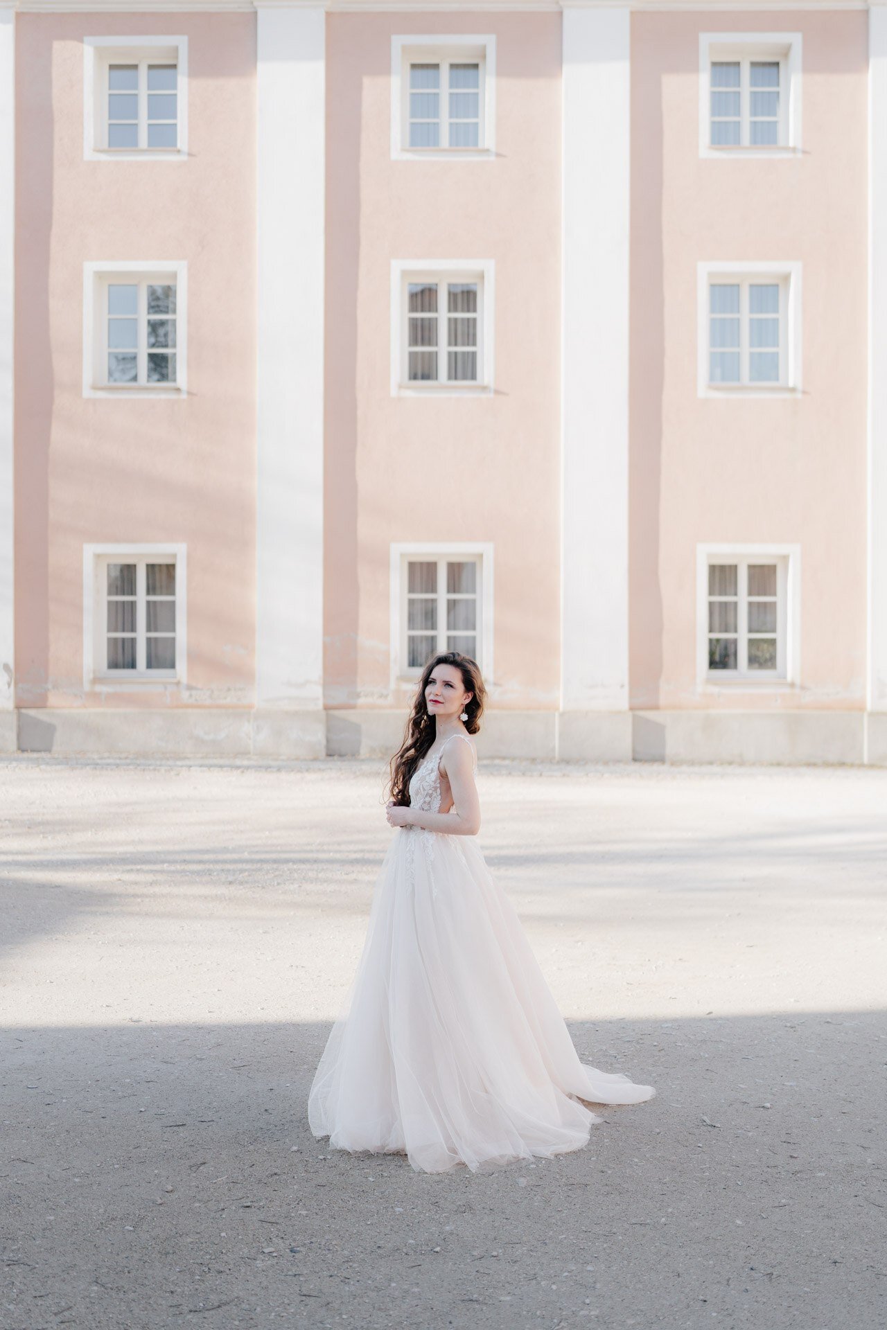 Eine Frau in einem fließenden weißen Kleid steht vor einem blassrosa Gebäude mit sechs weiß umrandeten Fenstern, das in sanftes Tageslicht getaucht ist - eine bezaubernde Szene, die jeder Hochzeitsfotograf Pforzheim perfekt festhalten kann.