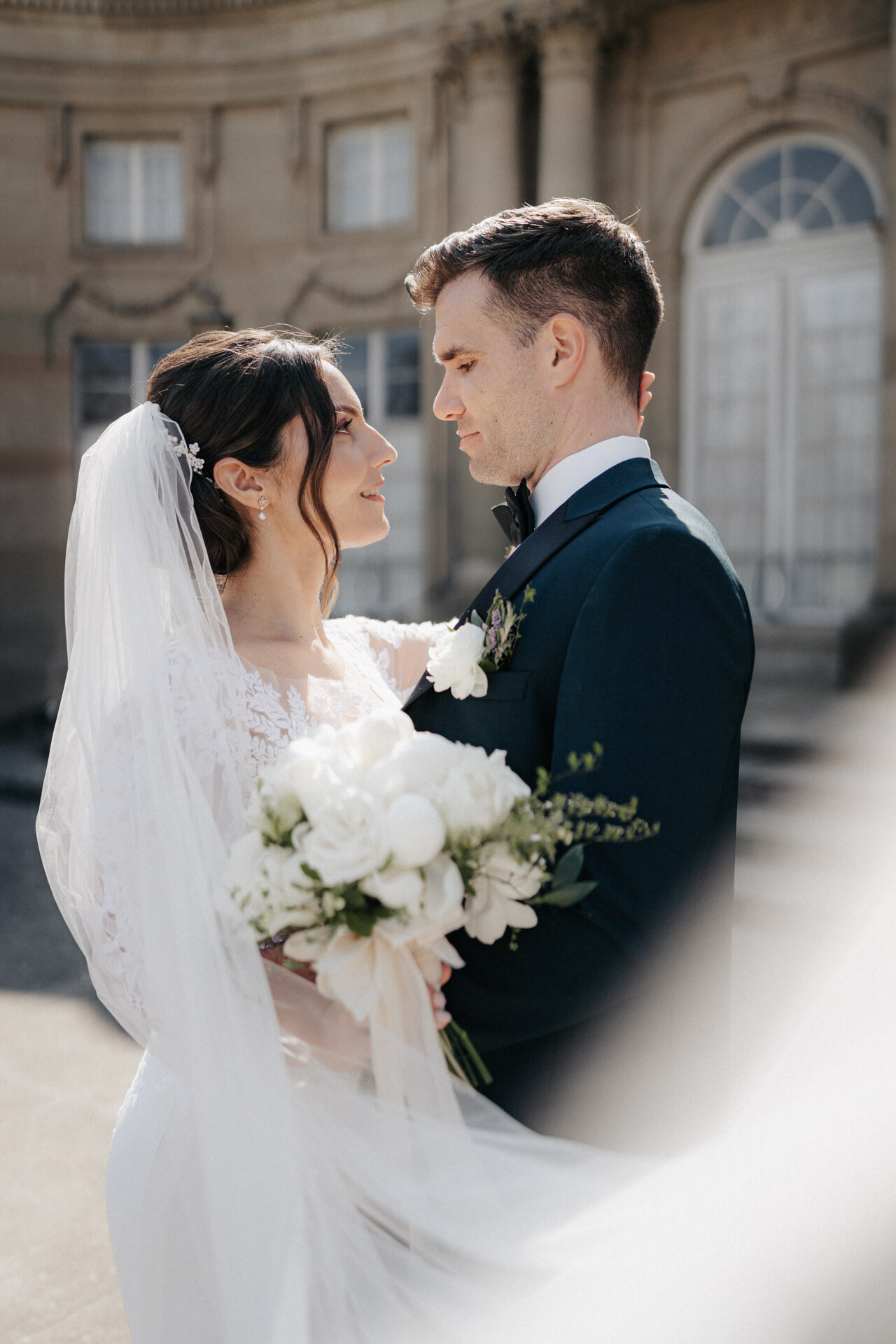 Eine Braut und ein Bräutigam stehen im Freien eng beieinander und blicken sich in die Augen, während das Sonnenlicht ihre Gesichter beleuchtet. Die von einem Hochzeitsfotografen aus Pforzheim aufgenommene Braut hält einen weißen Blumenstrauß und trägt ein Spitzenkleid neben ihrem Bräutigam in einem dunklen Anzug.