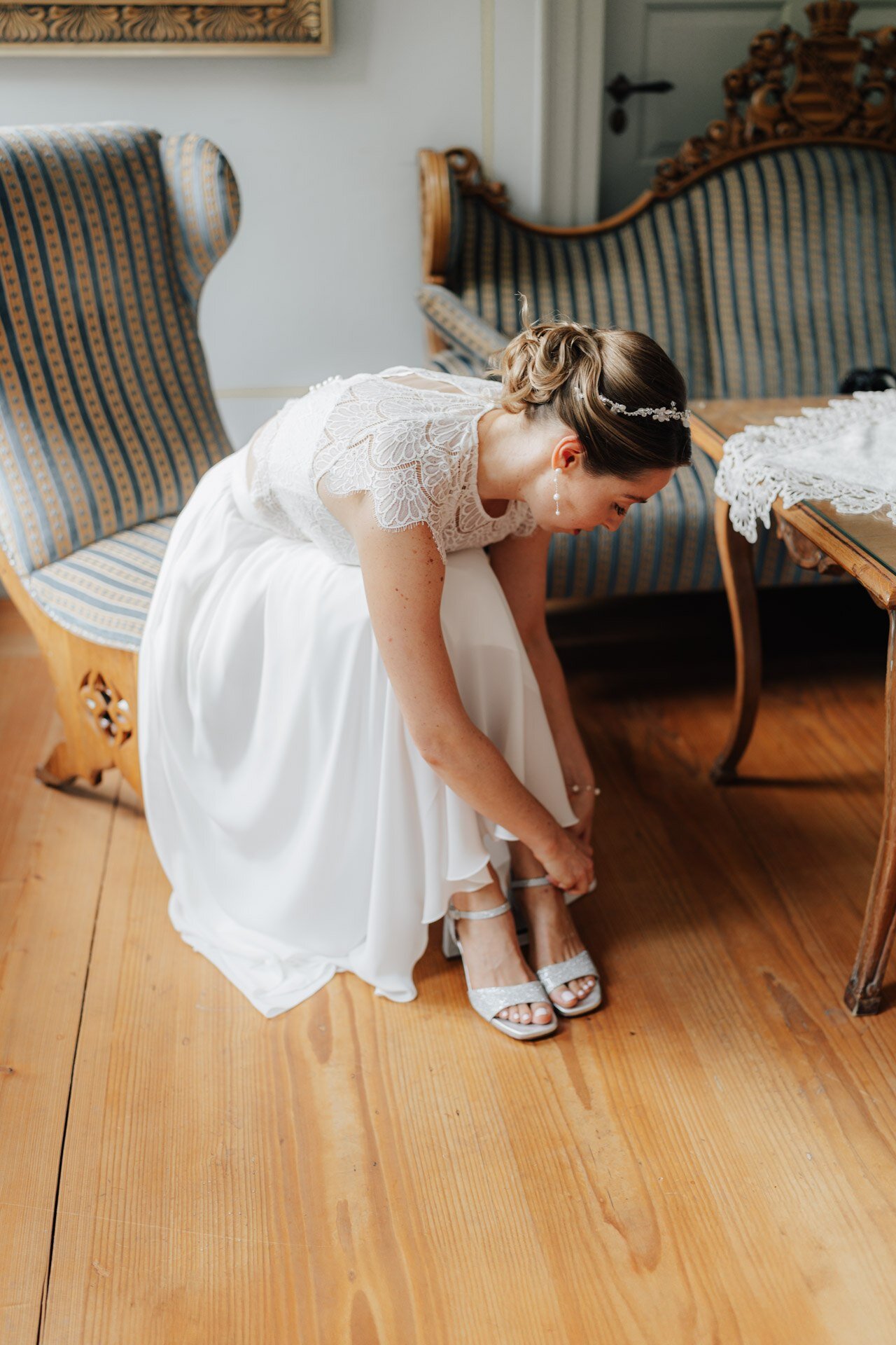 Eine Braut in einem weißen Spitzenkleid sitzt auf einem Vintage-Stuhl und lehnt sich vor, um ihre Sandale zu schließen. Im Hintergrund sind Holzböden und klassische Möbel zu sehen - wunderschön eingefangen von Hochzeitsfotograf Pforzheim.