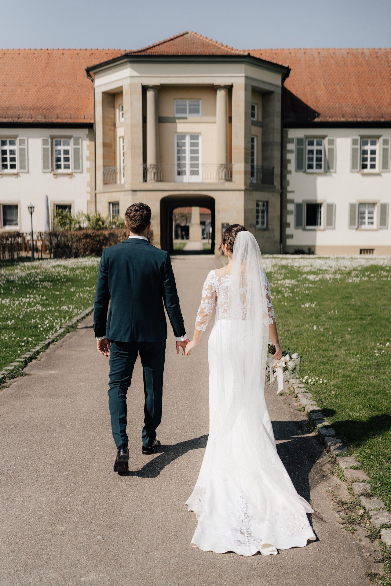 Braut und Bräutigam schreiten an einem sonnigen Tag Hand in Hand auf ein großes, elegantes Gebäude zu - ein perfekter Moment, eingefangen von einem Hochzeitsfotografen Pforzheim, mit der Braut in einem weißen Kleid und Schleier und dem Bräutigam in einem dunklen Anzug.