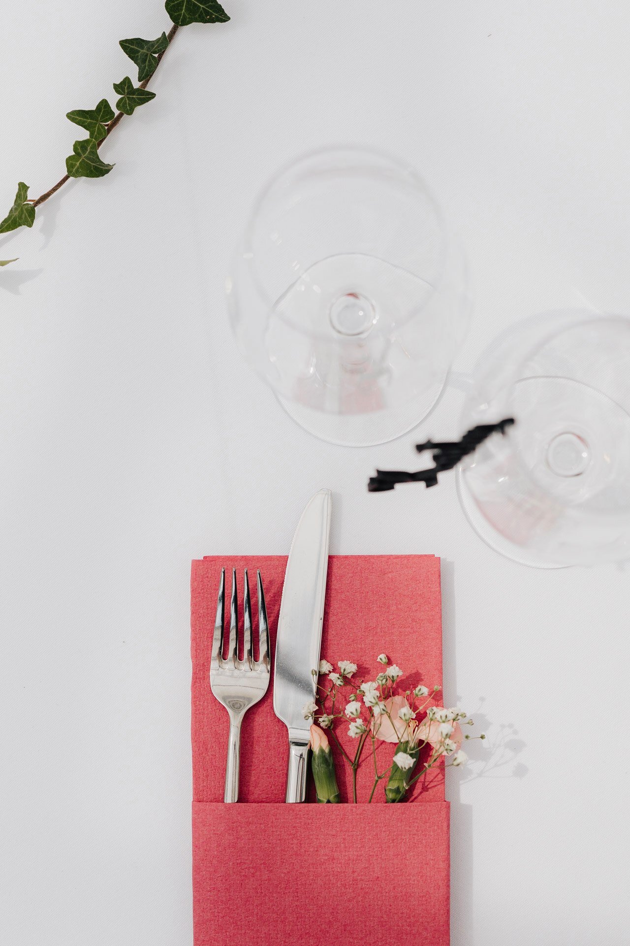 Eine Gabel und ein Messer liegen auf einer roten Serviette mit kleinen weißen Blumen daneben. Zwei leere Weingläser und eine grüne Efeuranke sind auf einem weißen Hintergrund zu sehen - die perfekte Inspiration für einen Hochzeitsfotografen Pforzheim.