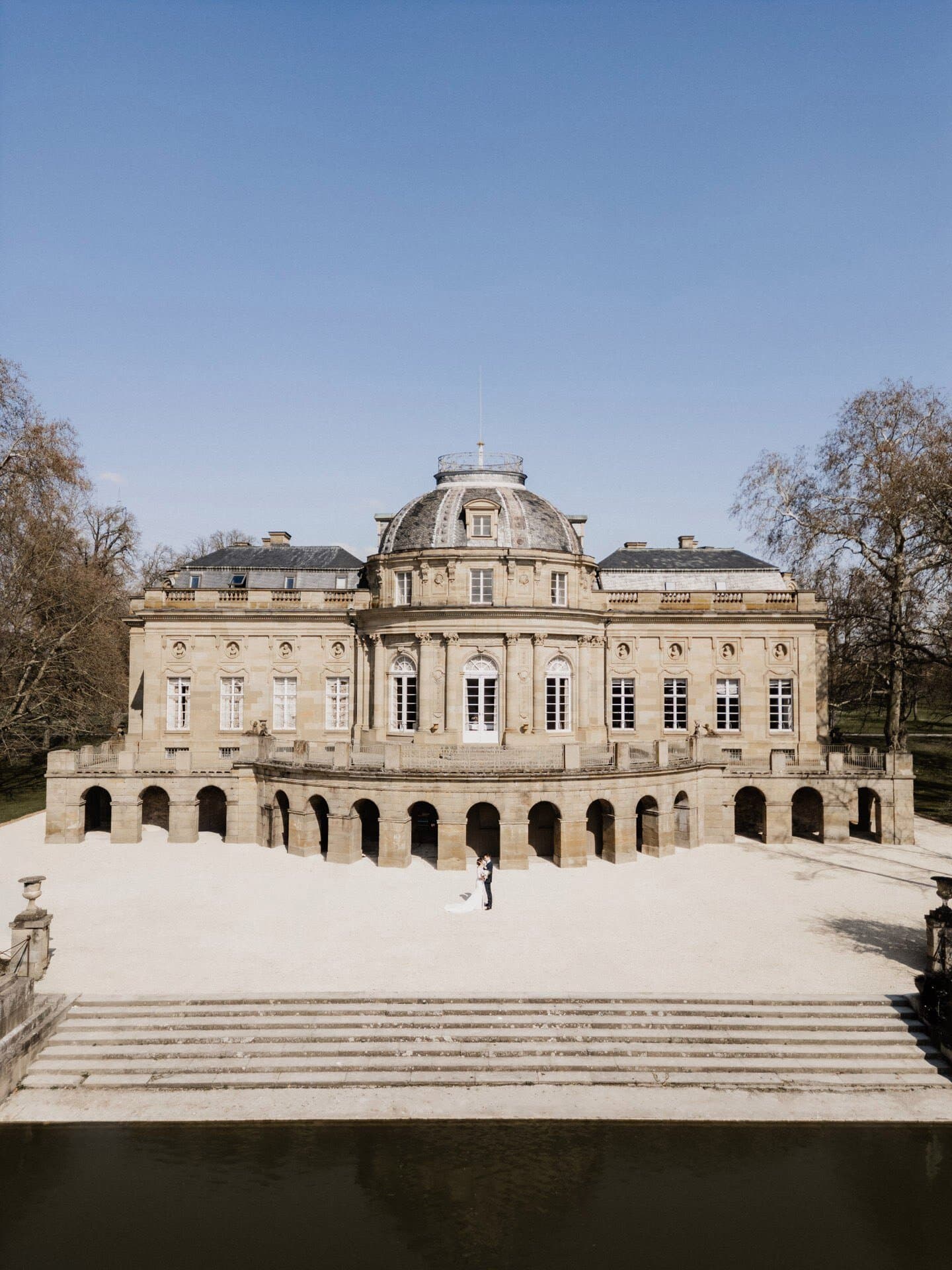 Ein großes historisches Steinschloss mit symmetrischen Fenstern und einem Kuppeldach steht unter einem klaren blauen Himmel; ein Paar in Hochzeitskleidung posiert im Innenhof, wunderschön eingefangen von einem Hochzeitsfotograf Stuttgart.