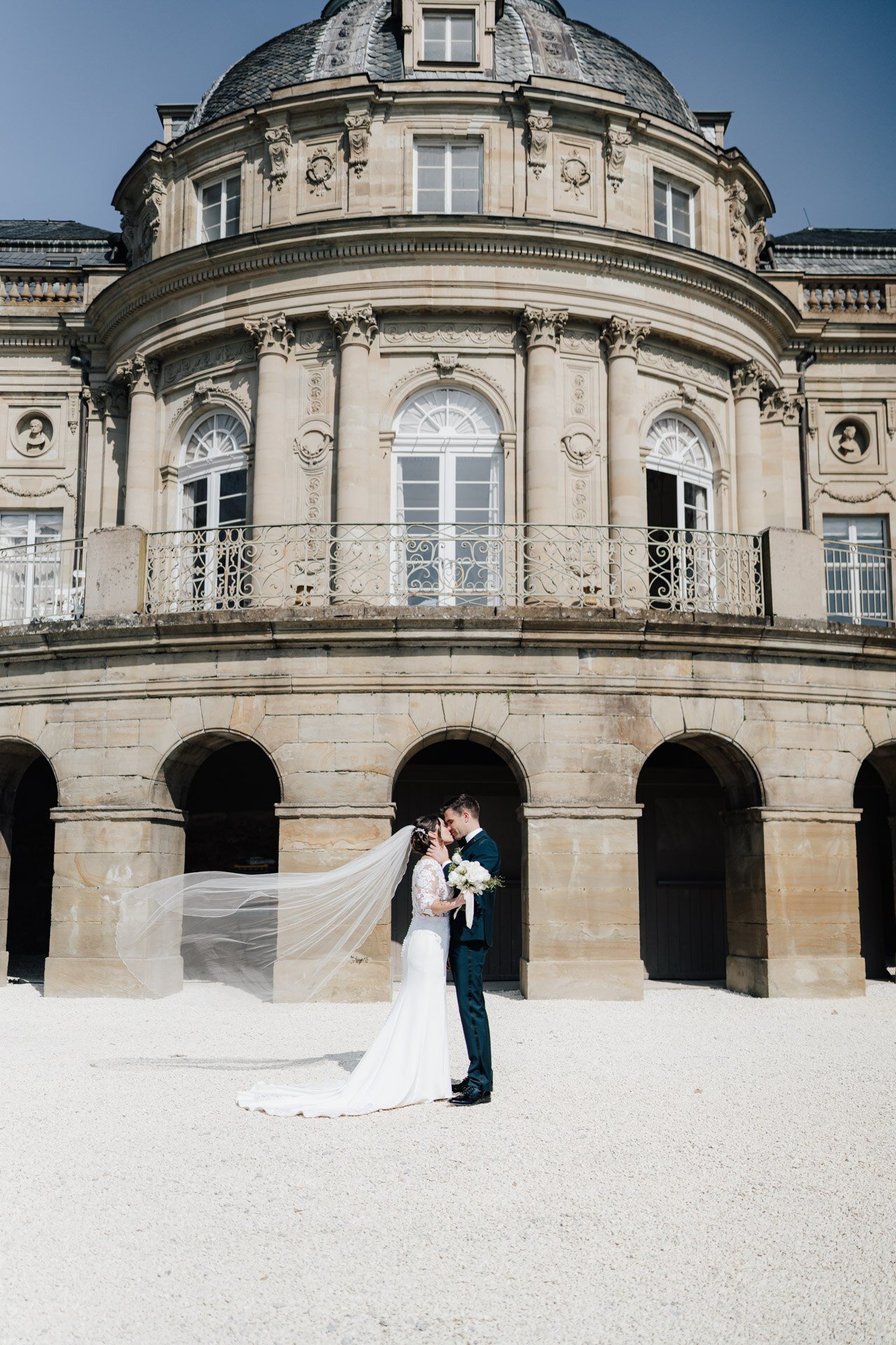 Braut und Bräutigam stehen sich umarmend vor einem prächtigen, historischen Steingebäude in Tübingen gegenüber. Der Schleier der Braut weht im Wind und die beiden werden von hellem Sonnenlicht umrahmt, aufgenommen von einem Hochzeitsfotograf Tübingen.
