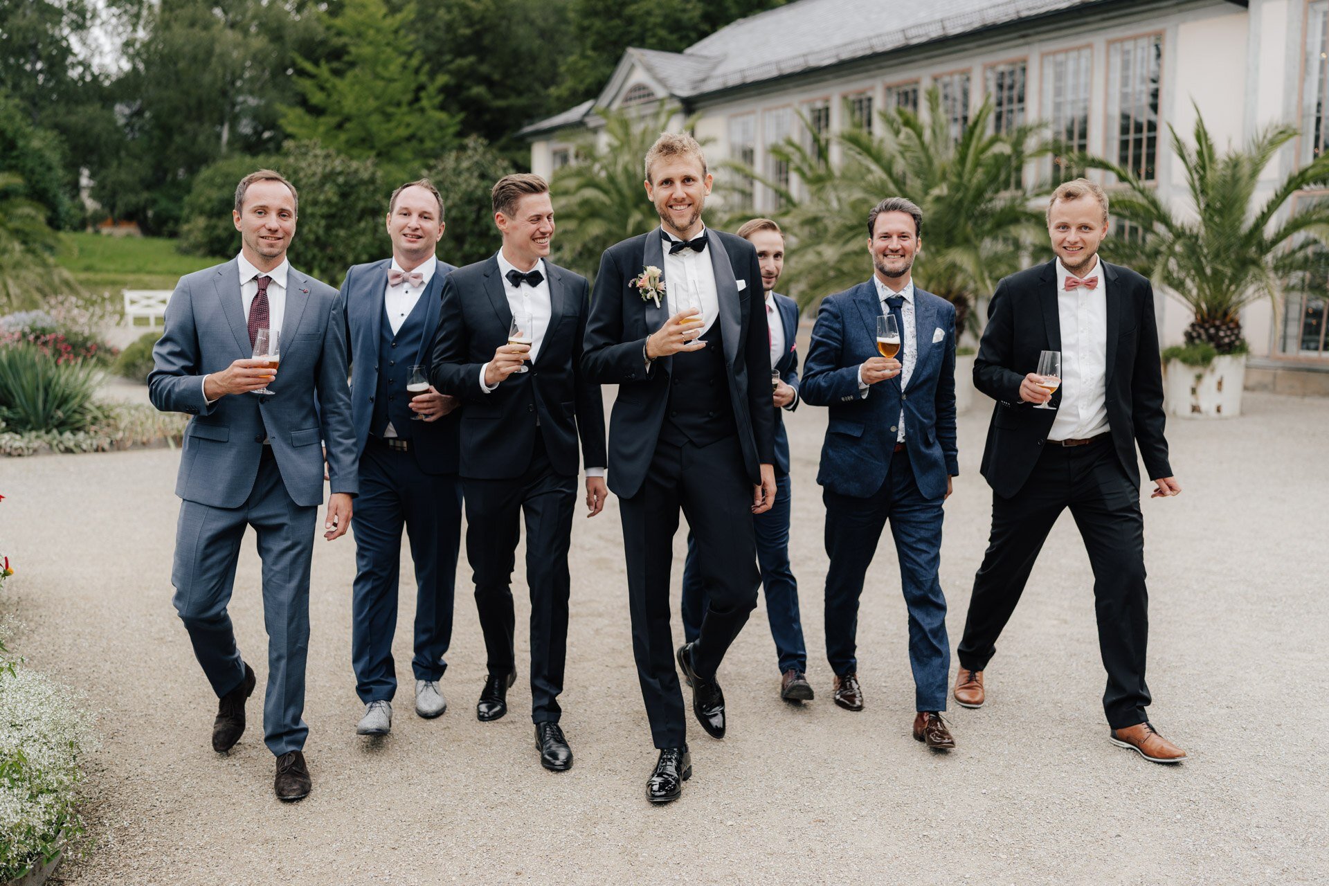 Eine Gruppe von sieben Männern in Anzügen, lächelnd und mit Getränken in der Hand, geht gemeinsam im Freien in der Nähe eines Gartens und eines großen Glasgebäudes spazieren, brillant eingefangen von einem Hochzeitsfotografen aus Tübingen - was auf ein feierliches Ereignis wie eine Hochzeit hindeutet.