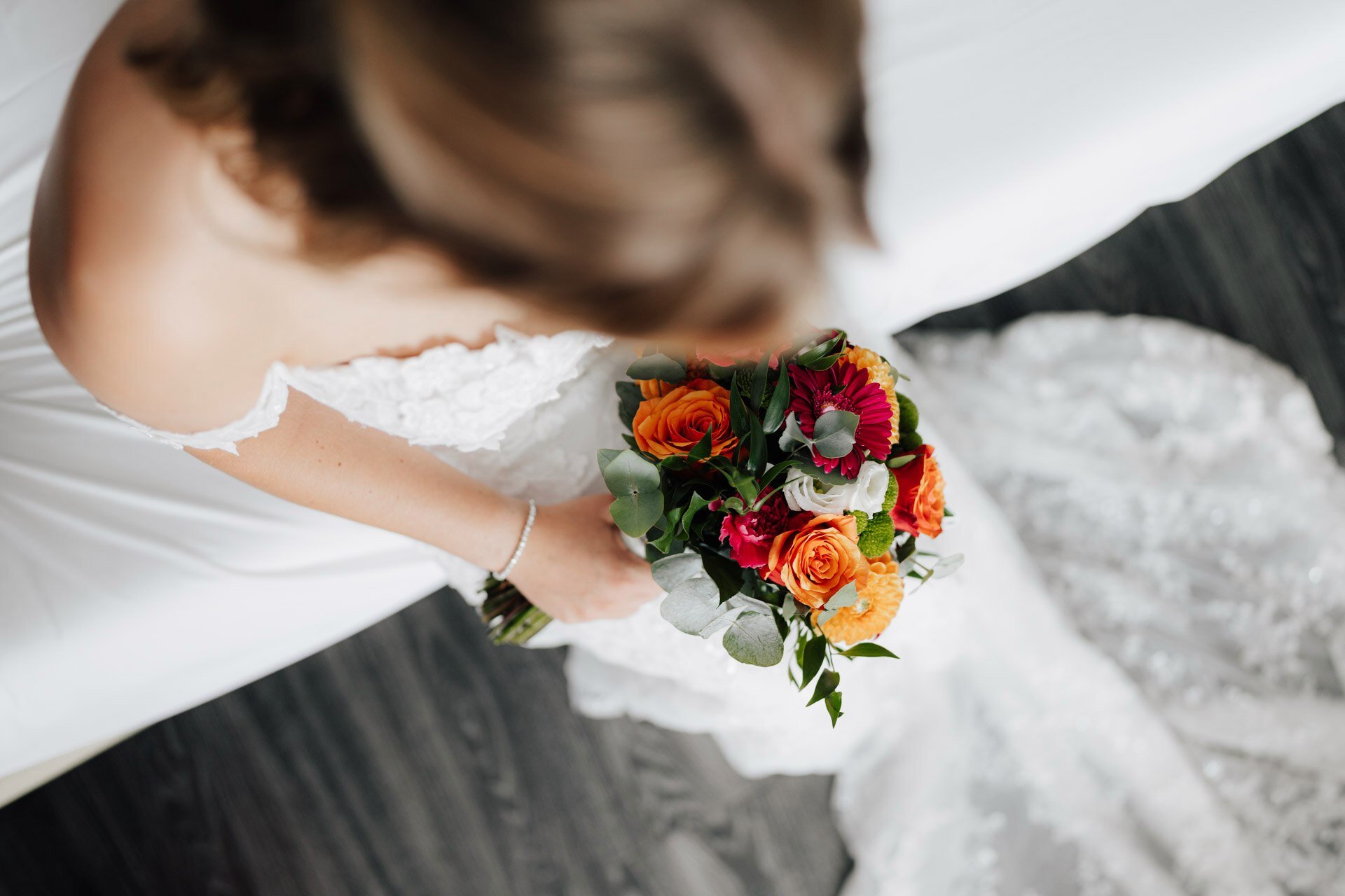 Eine Braut in einem weißen Spitzenhochzeitskleid hält einen leuchtenden Strauß aus orangefarbenen und rosafarbenen Blumen, fotografiert von einem Hochzeitsfotografen aus Tübingen von oben, wobei ihr Gesicht leicht unscharf ist. Sie sitzt auf einer weißen Fläche mit einem dunklen Boden darunter.